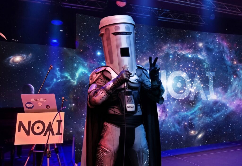 Count Binface wears a silver cape and large silver trashcan on his head while he speaks at a generated image of a podium with NOAI written on it